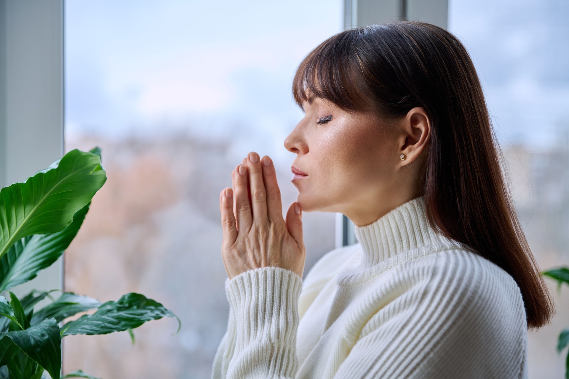 Middle aged woman folded hands in prayer meditation at home near window